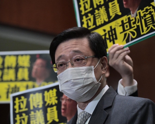 李家超在立法會見傳媒時，民主派議員舉示威標語。