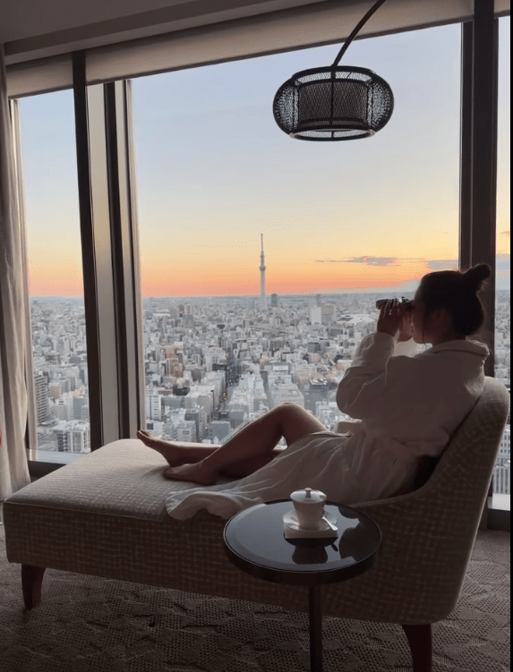 黃碧蓮在房間內身穿浴袍，手持望遠鏡欣賞東京景色。