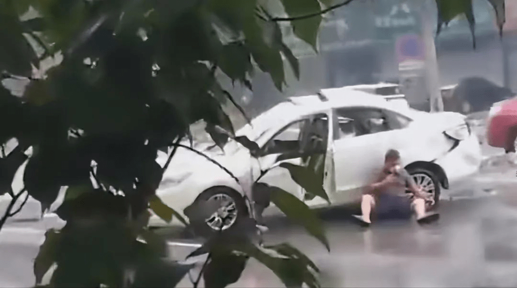 网片显示，一辆白色汽车受损严重，一名男子背靠汽车坐在地上。
