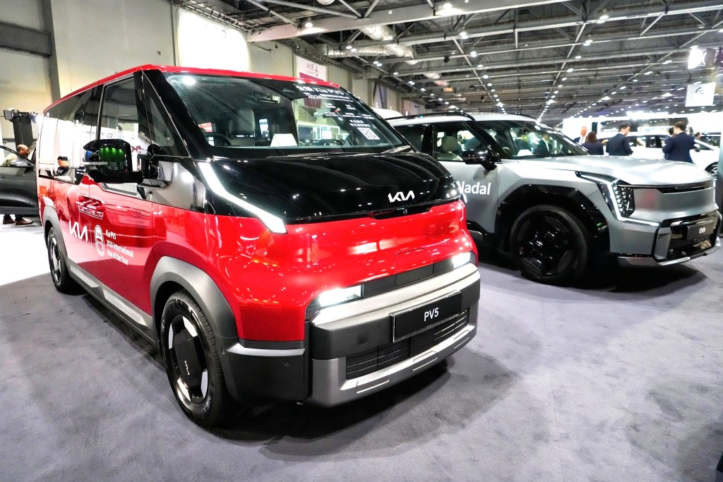 A red and black Kia PV5 electric MPV with a sleek, modern design is displayed alongside a silver SUV at an indoor auto exhibition, with people observing in the background.