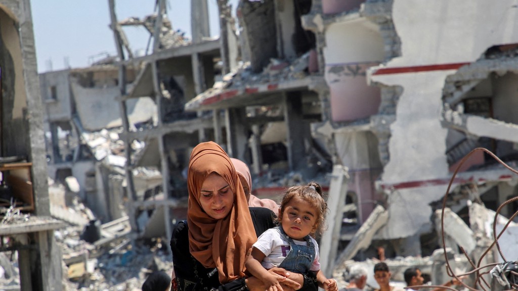 以軍攻擊加沙地帶多區，圖為汗尤尼斯一名母親抱着女兒走過碎成瓦礫的民房。 路透社
