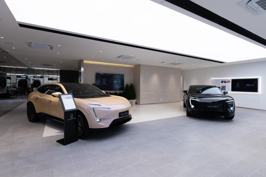 Two AVATR electric SUVs are displayed in a modern, brightly lit showroom, with the cream-colored model on the left featuring a sleek design and a digital information stand, while the black model on the right showcases a sporty appearance; the showroom has minimalist decor with digital screens on the wall and a reflective glass partition in the background.
