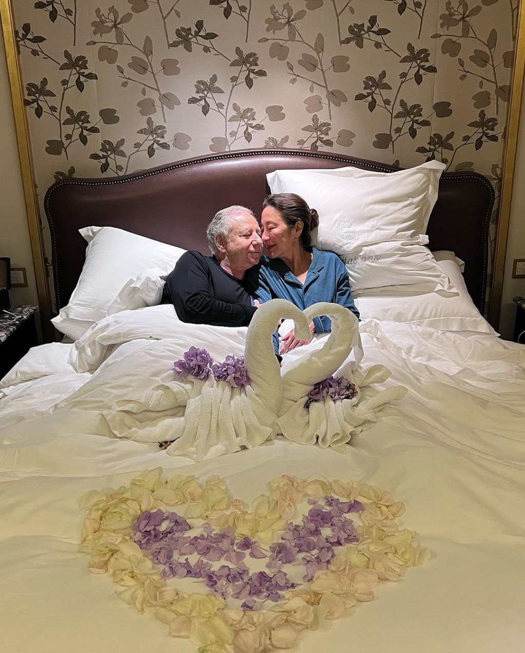 一對新人在以「心形毛巾天鵝」佈置及鋪滿花瓣的床上依偎。