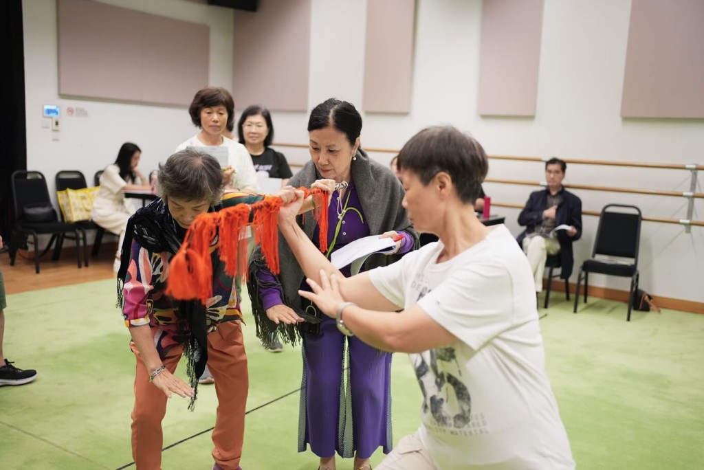 一身紫色長袖打扮的汪明荃先跟該粵劇演員對稿，然後拿起道具排練。