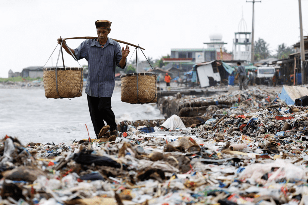 印尼萬丹省班德蘭攝政區，在直落漁村的海灘上，一名背著籃子的老人走過成堆的垃圾，其中大部分是塑膠和生活垃圾。2024 年 3 月 15 日。路透社路透社