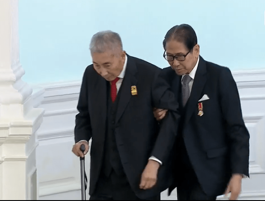 梁智仁在胞兄梁智鴻的攙扶上台受勳。港台FB影片截圖