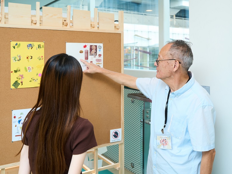 ET(右)於「生命旅程展覽」中向參觀者介紹長者的藝術作品。(領展提供)