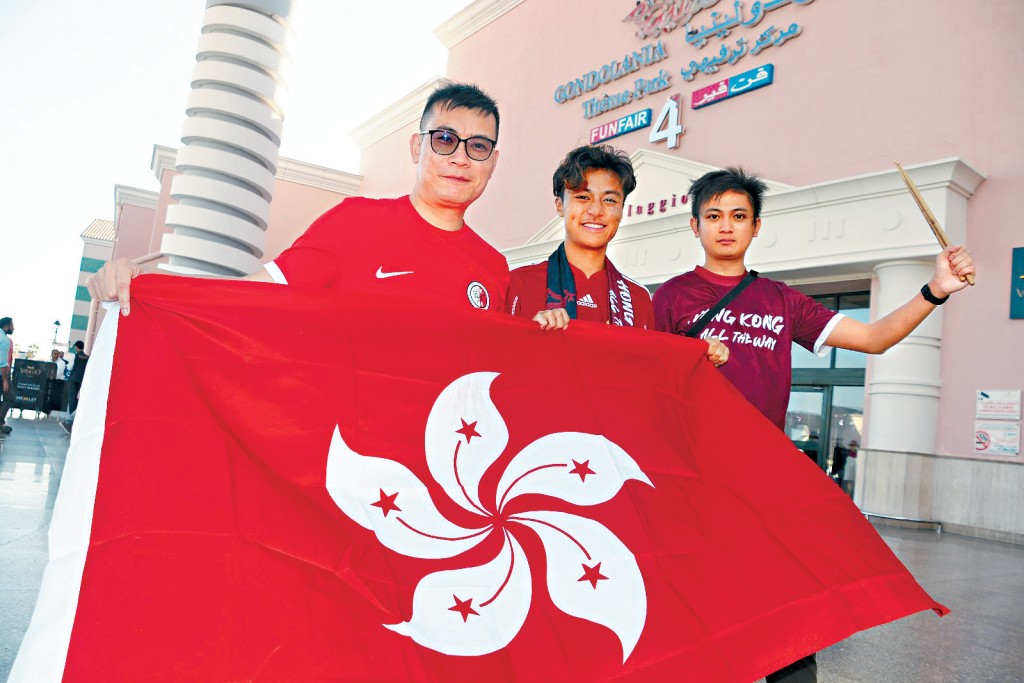 三位港足球迷(左起)崔先生、Aidan及恆仔於哈利法國際球場附近,舉起旗幟造勢。