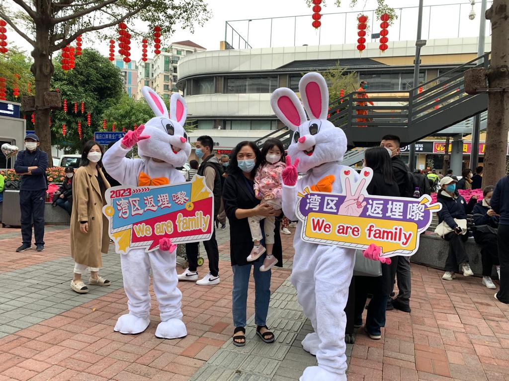 在福田口岸外，有兔子打扮的工作人員手持「灣區返埋嚟 We are family」字樣的紙牌，有市民上前打卡。（常彧璠攝）