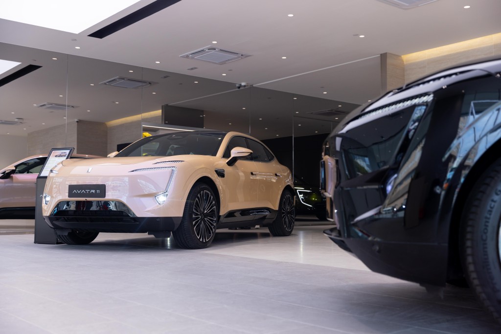 A white AVATR 11 electric SUV with a sleek design and dual screens in the front, displayed alongside other luxury vehicles at the launch event in a modern showroom.