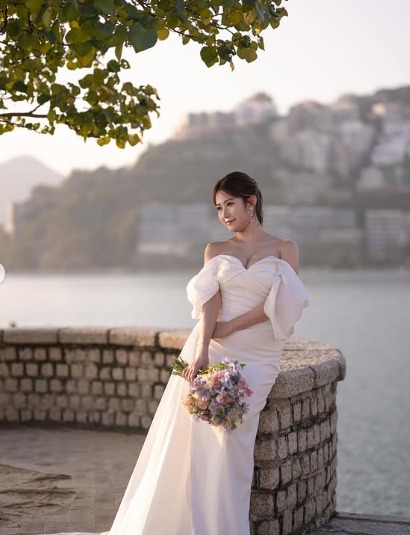 黃婧靈化上淡妝,梳著中分低馬尾的髮型,身穿白色一字領露膊低胸緞面婚紗,亦手持大束紫紅色鮮花在沙灘邊留影。 黃婧靈化上淡妝,梳著中分低馬尾的髮型,身穿白色一字領露膊低胸緞面婚紗,亦手持大束紫紅色鮮花在沙灘邊留影。
