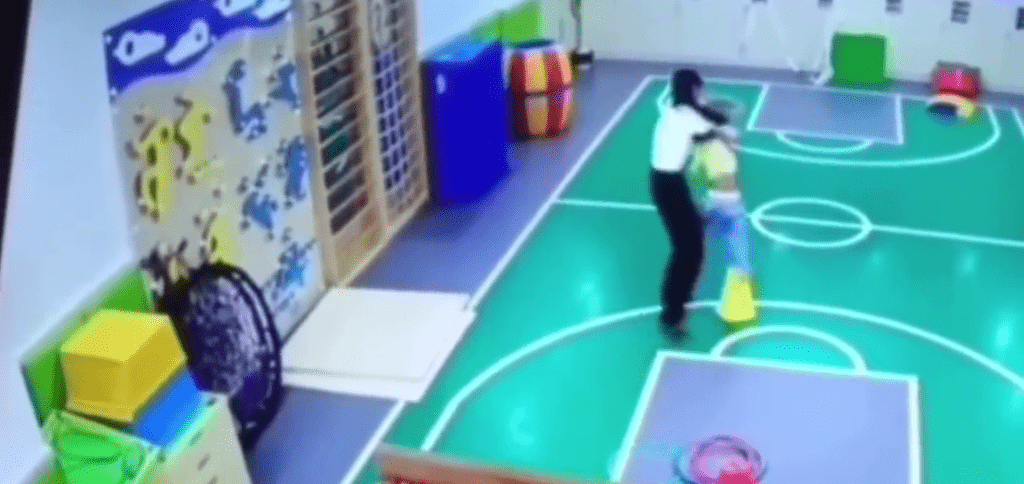 老师强行将幼童拉起来，让他站在橡胶雪糕筒上。