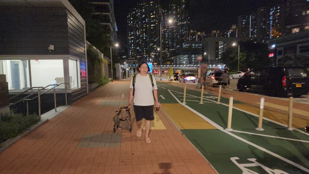 關女身穿白色T恤及黑色短褲，背上背囊並拖着手拉車，手持飲品步出大樓。黎志偉攝