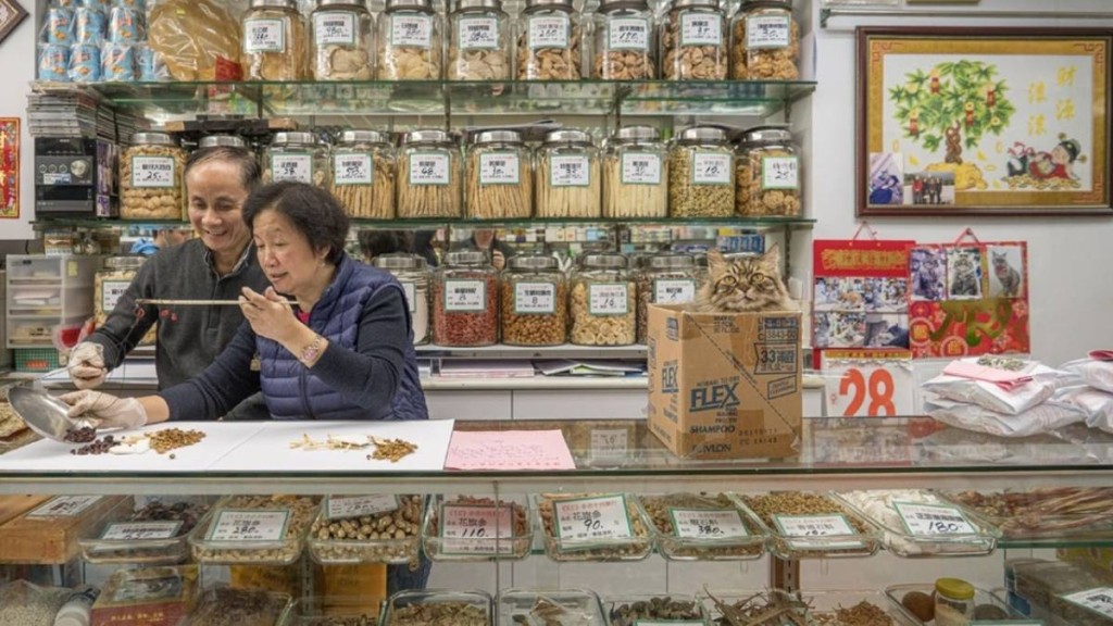 香港中藥行東主秤量藥材，旁邊紙皮箱躲着一隻褐貓。Blue Lotus Gallery提供