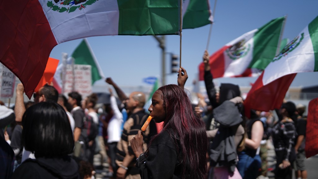 洛杉矶示威|一名示威者在挥舞墨西哥国旗的人群中吃冰棒降温。 美联社 洛杉矶示威|一名示威者在挥舞墨西哥国旗的人群中吃冰棒降温。 美联社