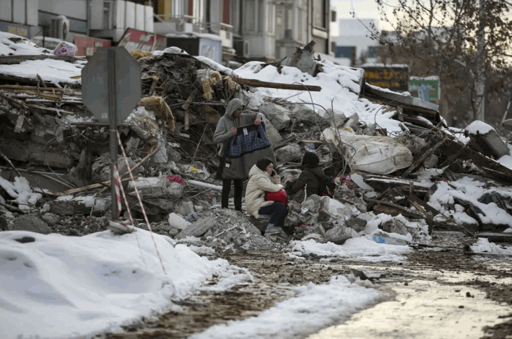 人們坐在土耳其馬拉蒂亞一座倒塌的建築物旁。搜救隊和援助正湧入土耳其和敘利亞，救援人員在冰凍溫度下挖掘被地震夷為平地的建築物殘骸。AP
