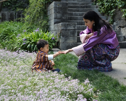 小王子坐在皇宮花園草地上,伸手觸碰媽媽佩瑪的手。