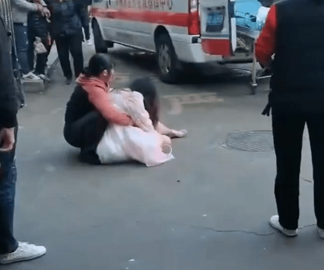 一名女生瘫坐在地上，衣服、地面疑似有血迹，身旁有一名妇女正抱著她。