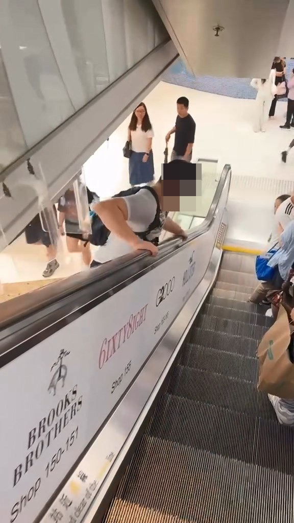 男子翻越玻璃欄杆,爬落扶手電梯中間的銀色蓋板,之後沿著蓋板慢慢爬行到下一層。Threads@showerrrjun片段截圖 男子翻越玻璃欄杆,爬落扶手電梯中間的銀色蓋板,之後沿著蓋板慢慢爬行到下一層。Threads@showerrrjun片段截圖