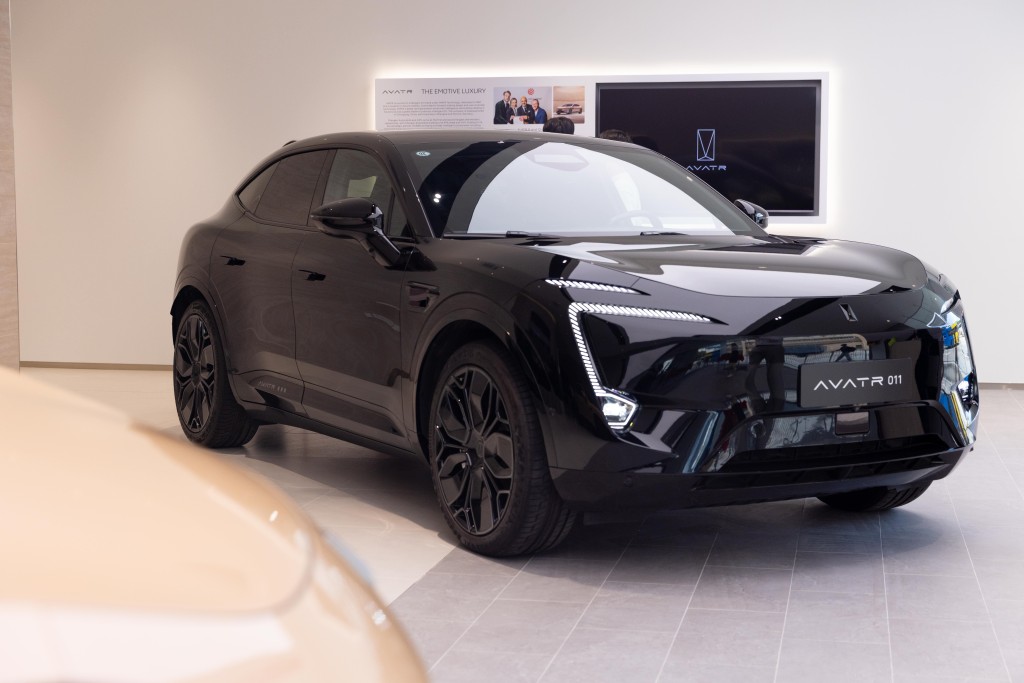 A sleek black AVATR 011 electric SUV with a modern design and distinctive front lighting is displayed in a showroom with a white wall and a digital screen behind it.