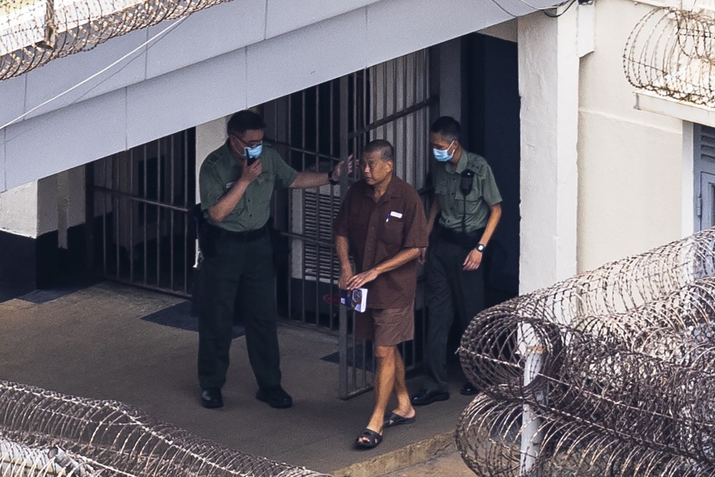 黎智英身穿傳統啡色囚服，穿着拖鞋，手拿一本書，由兩名懲教人員陪同外出。AP