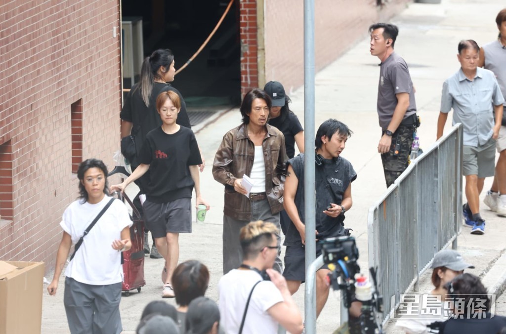 手握劇本的黃子華與幕後人員並肩穿過殯儀館旁巷道，準備前往下一拍攝點。